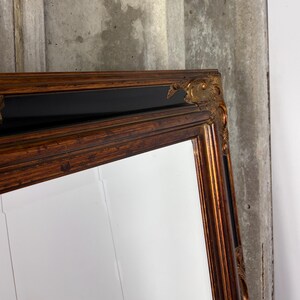 May include: A rectangular wall mirror featuring a wide, ornate frame with a dark wood-toned finish and black accents. The corners of the frame are decorated with raised, gilded gold-colored carvings that add texture and detail to the structure.