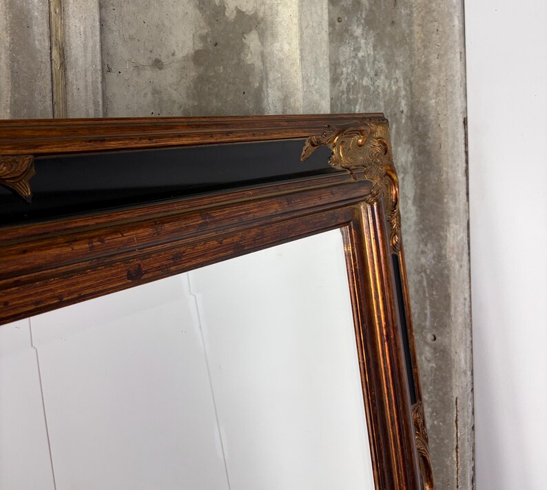 May include: A rectangular wall mirror featuring a wide, ornate frame with a dark wood-toned finish and black accents. The corners of the frame are decorated with raised, gilded gold-colored carvings that add texture and detail to the structure.