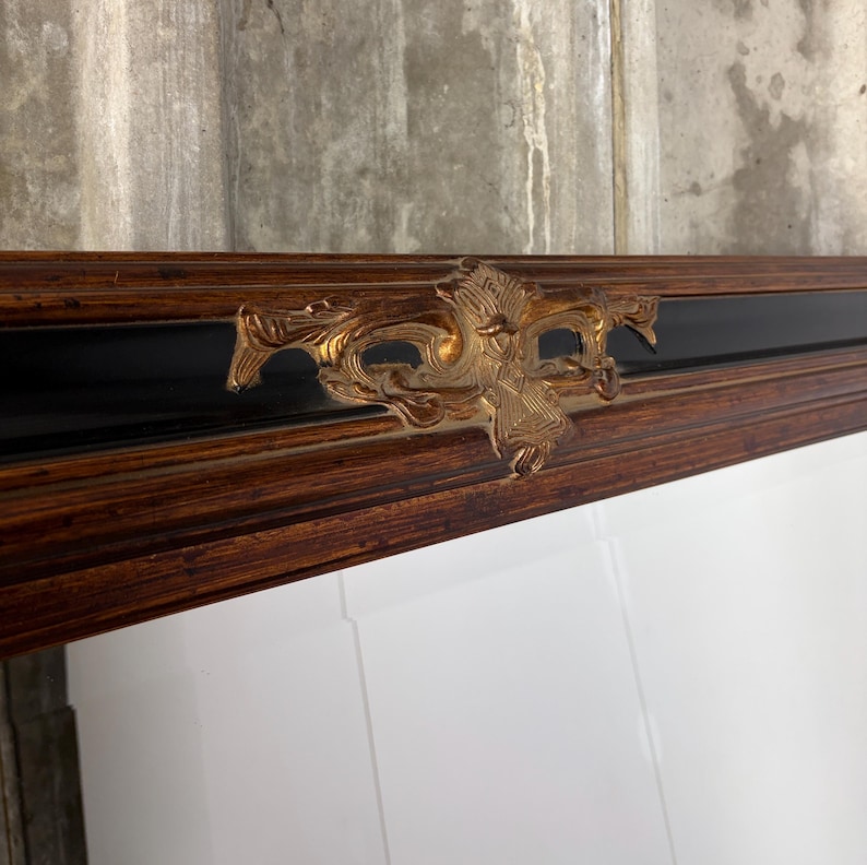 May include: A rectangular wall mirror featuring a dark brown wood grain frame with a smooth black inner border. A prominent, ornate gold-colored metal decorative accent with intricate scrollwork and relief detailing is centered on the top frame.