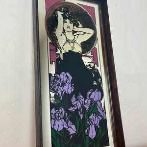 May include: Art Nouveau style print in a dark wood frame. The print features a woman with her arms raised, surrounded by a circular design. Below the woman are purple iris flowers against a black background. The frame is a dark brown color.