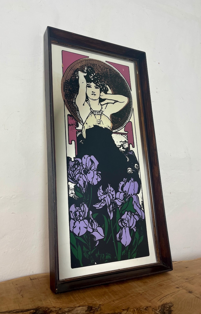 May include: Art Nouveau style print in a dark wood frame. The print features a woman with her arms raised, surrounded by a circular design. Below the woman are purple iris flowers against a black background. The frame is a dark brown color.