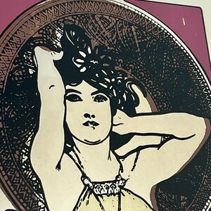 May include: Art Nouveau style print featuring a woman with dark hair, framed by an ornate circular border. The woman is depicted in a cream-colored dress, with her arm raised. The background is a mix of pink and gold tones.