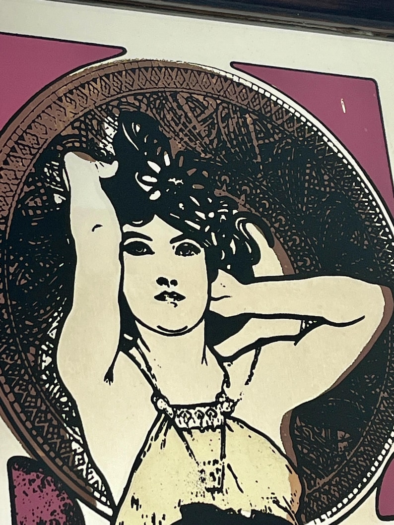 May include: Art Nouveau style print featuring a woman with dark hair, framed by an ornate circular border. The woman is depicted in a cream-colored dress, with her arm raised. The background is a mix of pink and gold tones.