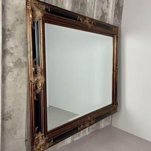 May include: A large rectangular wall mirror featuring a wide, ornate frame with a combination of dark wood tones and black accents. The corners and center edges of the frame are decorated with intricate, raised gold-colored floral carvings.