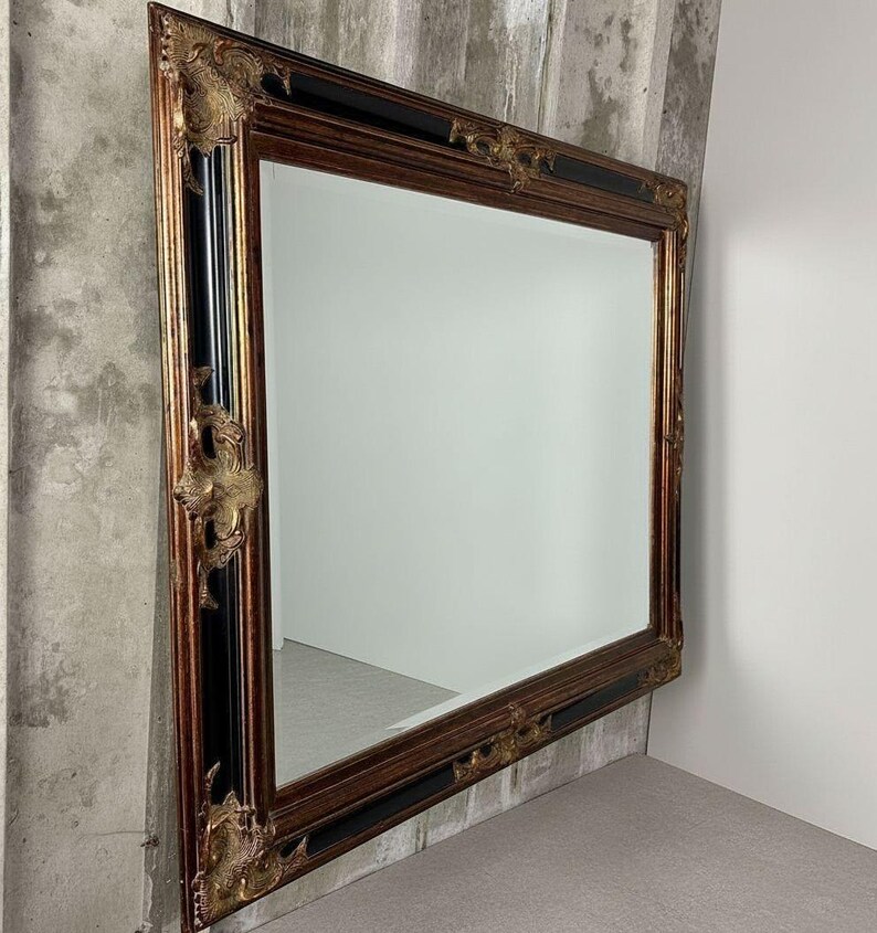 May include: A large rectangular wall mirror featuring a wide, ornate frame with a combination of dark wood tones and black accents. The corners and center edges of the frame are decorated with intricate, raised gold-colored floral carvings.