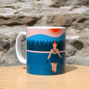 May include: A white ceramic mug with a blue, pink, and orange illustration of a person in a swimsuit standing in front of a lake and forest. The sun is setting behind the person.