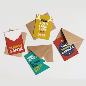 May include: A collection of Christmas greeting cards with brown envelopes. The cards feature festive text in various colors, including red, yellow, and teal. The phrases include "SLAINTE SANTA", "FOR AULD LANG SYNE MA DEER", "SUCH BONNIE BAUBLES", and "HAVE YERSEL A BONNIE WEE CHRIST MAS".