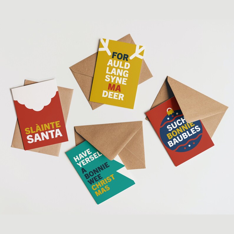 May include: A collection of Christmas greeting cards with brown envelopes. The cards feature festive text in various colors, including red, yellow, and teal. The phrases include "SLAINTE SANTA", "FOR AULD LANG SYNE MA DEER", "SUCH BONNIE BAUBLES", and "HAVE YERSEL A BONNIE WEE CHRIST MAS".