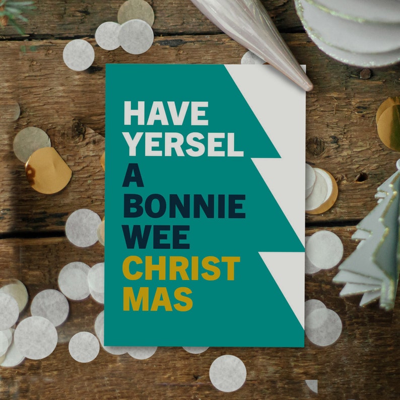 May include: A Christmas card with the text "HAVE YERSEL A BONNIE WEE CHRISTMAS" in white and gold lettering on a teal green background. The card is decorated with a Christmas tree design and sits on a wooden surface with confetti.