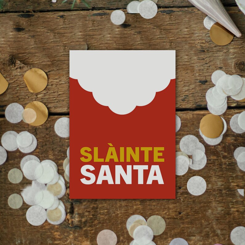 May include: A festive greeting card with a red background and a white cloud-shaped design at the top. The words "SL&Agrave;INTE SANTA" are printed in gold and white. The card is surrounded by confetti on a wooden surface.