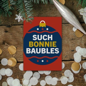 May include: A red greeting card with the text "SUCH BONNIE BAUBLES" in white and gold lettering, inside a blue and red ornament design. The card is on a wooden surface with confetti and Christmas decorations.