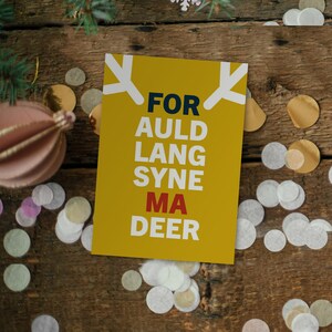 May include: A yellow greeting card with the text "FOR AULD LANG SYNE MA DEER" in navy blue and red. The card has white antler-like shapes at the top. The card is on a wooden surface with confetti and a pink ornament.