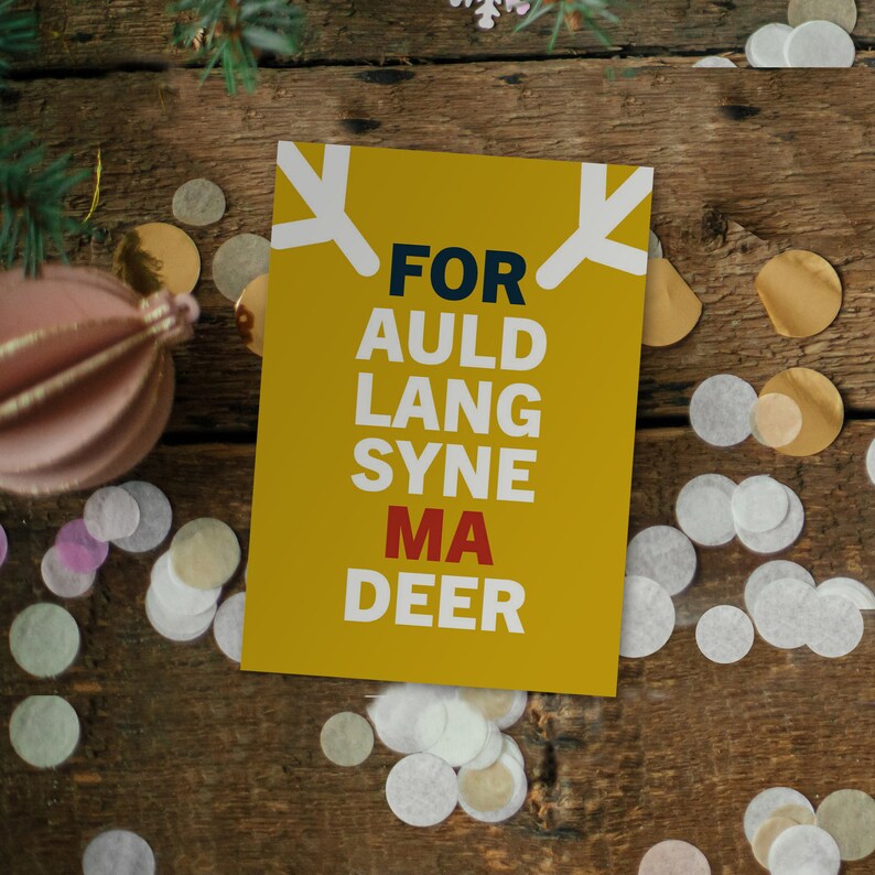 May include: A yellow greeting card with the text "FOR AULD LANG SYNE MA DEER" in navy blue and red. The card has white antler-like shapes at the top. The card is on a wooden surface with confetti and a pink ornament.