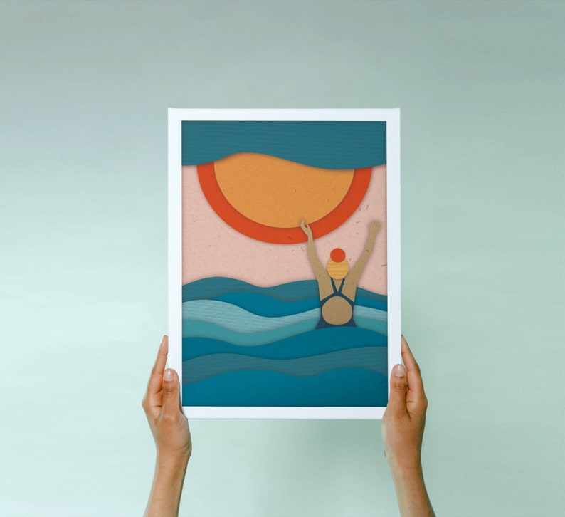 May include: A framed illustration of a person swimming in the ocean with a large orange sun in the background. The illustration is made with paper cutouts and has a layered effect.