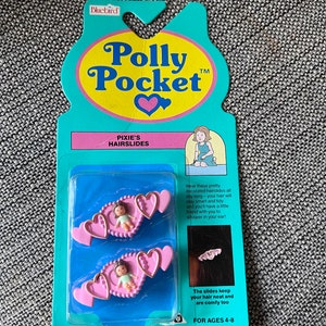 May include: A package of two pink heart-shaped hair slides with a small doll figurine in the center of each. The package is blue and yellow and says "Polly Pocket" and "Pixie's Hairslides".