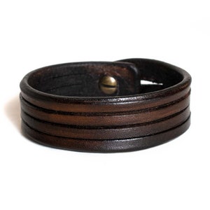 Pode incluir: Um bracelete de couro marrom com várias camadas e um fecho de metal.