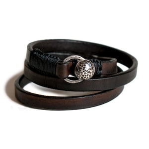 Könnte beinhalten: Ein dunkelbraunes Lederarmband zum Wickeln mit einer silberfarbenen, strukturierten Perle und einem Verschluss. Das Armband wird mehrmals um das Handgelenk gewickelt. Ein schwarzes Kordeldetail ist in der Nähe des Verschlusses um das Leder gewickelt.