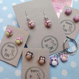 May include: Handmade owl-themed jewelry and accessories. The image features pink, brown, and purple owl earrings, stud earrings, a keychain, and a necklace pendant. Each item is attached to a kraft paper card with the text "Handmade by Vani" and a fox logo.
