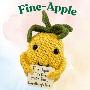 Könnte beinhalten: Ein gehäkelter gelber Apfel mit grünem Stiel und Blättern, der ein Schild mit der Aufschrift "Fine-Apple It's fine. You're fine. Everything's fine." hält.
