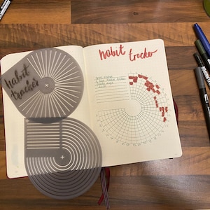 Peut inclure: Une page de suivi des habitudes dans un journal. La page est divisée en sections avec une grille circulaire et une liste d'habitudes. Les habitudes sont marquées de points rouges. La page est intitulée "habit tracker" en écriture cursive. Il y a deux pochoirs sur la page, l'un avec un motif en forme de soleil et l'autre avec des cercles concentriques.