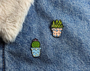 Puede incluir: Dos pines de esmalte con forma de suculentas en macetas. Un pin presenta una suculenta verde en una maceta azul con lunares blancos y caras sonrientes. El otro pin tiene una suculenta verde en una maceta rosa con lunares azules y caras sonrientes.