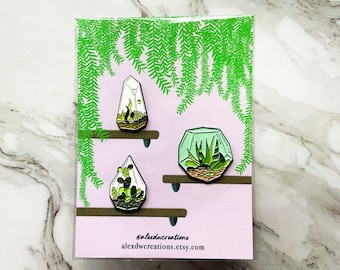 May include: Three terrarium enamel pins displayed on a pink card with green fern illustrations. The pins depict geometric glass shapes with miniature succulent plants. The card includes the text "@alexdwcreations alexdwcreations.etsy.com".
