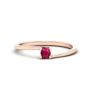 May include: A delicate rose gold ring with a single, round, red gemstone set in a prong setting. The band is thin and has a curved design.