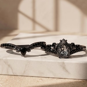 Op de afbeelding: Twee zwarte ringen. Eén ring heeft een peervormige, grijze edelsteen met zwarte accenten. De andere ring heeft een gebogen band met een zwarte druppelvormige steen en zwarte bladachtige details. Beide ringen liggen op een wit marmeren oppervlak.