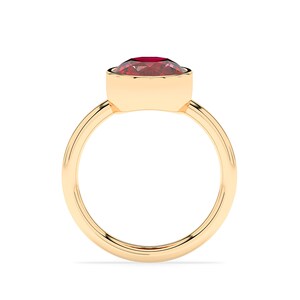 Könnte beinhalten: Ein goldener Ring mit einem großen, oval geformten, tiefroten Edelstein. Der Edelstein ist in einer Zargenfassung gefasst, und das Band ist aus schlichtem, poliertem Gold. Der Ring wird vor einem weißen Hintergrund gezeigt, um sein elegantes Design hervorzuheben.