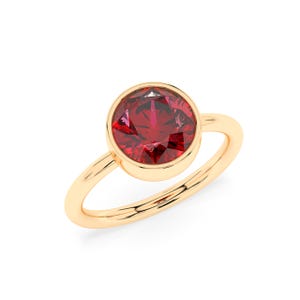May include: A gold ring with a large, round, faceted red gemstone. The gemstone is set in a bezel setting, and the band is a simple, polished gold. The ring is shown against a white backdrop.