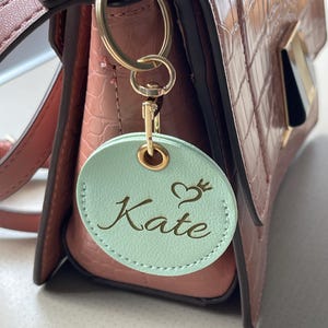 以下が含まれることがあります： ミントグリーンの丸いレザー製キーホルダー。筆記体で「Kate」という名前と、その上に小さなハートと王冠のアイコンが刻印されています。ゴールドトーンの金属製リングと留め具で、クロコダイル調のダスティピンクのハンドバッグに取り付けられています。