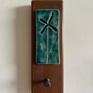 Puede incluir: Un colgador de pared de madera con una baldosa rectangular de cerámica turquesa con un diseño abstracto negro. La baldosa está montada sobre una base de madera marrón oscuro con un pequeño gancho de metal oscuro en la parte inferior.