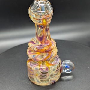 May include: A colorful glass pipe with a rounded bowl and a twisted, cylindrical body. The glass features swirls of yellow, orange, and purple. A small, clear glass handle is attached to the side of the base.