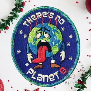May include: Embroidered patch with a distressed Earth graphic, tongue out, and the text "THERE'S NO PLANET B" in white. The patch is blue with white snowflakes and a green border. Christmas decorations surround the patch.