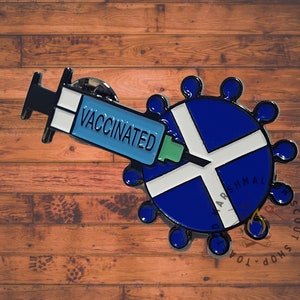 May include: A blue and white enamel pin featuring a syringe with the word "VACCINATED" and a circular design with a Scottish flag pattern. The pin is set against a wooden background.