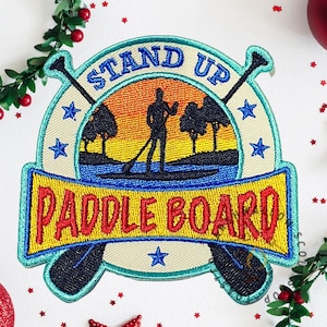 May include: Embroidered patch with the words "STAND UP PADDLE BOARD" in a circular design. The patch features a silhouette of a person paddleboarding on water, with trees and a sunset in the background. Blue stars and paddles are also included.