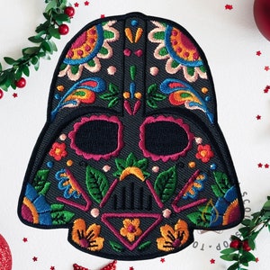 Op de afbeelding: Een zwarte patch in de vorm van Darth Vaders helm, versierd met kleurrijk bloemenborduurwerk. Het ontwerp bevat roze, gele, blauwe en groene elementen, waardoor een thema van de Dag van de Doden ontstaat. Feestelijke decoraties omringen de patch.
