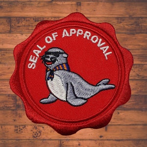 May include: Embroidered patch with a cartoon seal wearing sunglasses and a scarf, set against a red circular background with the words "SEAL OF APPROVAL" in white. The seal is grey with black sunglasses and a blue and orange scarf.