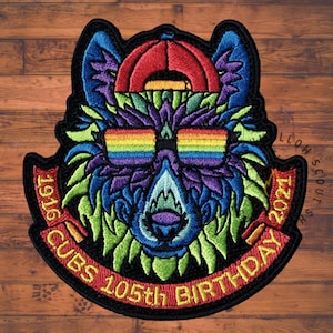 May include: Embroidered patch with a wolf head design, wearing a red cap and rainbow sunglasses. The patch has the text "CUBS 105th BIRTHDAY 1916-2021" in red and yellow. The wolf's fur is a mix of blue, purple, and green.