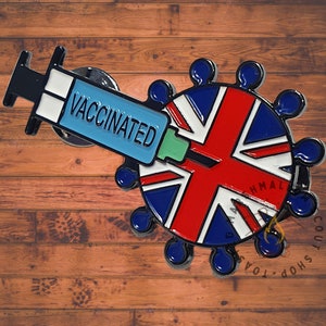 May include: A pin featuring a syringe with the word "VACCINATED" in blue, white, and black. The syringe is positioned over a circular design of the Union Jack flag, with blue and white accents. The pin is on a wooden surface.
