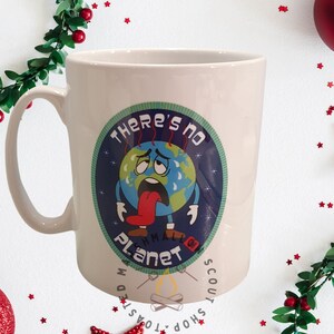 May include: White ceramic mug with a handle, featuring a distressed Earth graphic with the text "There's No Planet B". The Earth graphic is blue, green, and red, with a distressed look. The mug is surrounded by Christmas decorations.