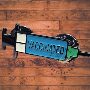 May include: A Vaccinated enamel pin in the shape of a syringe. The syringe has a black handle, a blue barrel with the word "VACCINATED" in white, and a green tip. The pin is on a wooden surface.