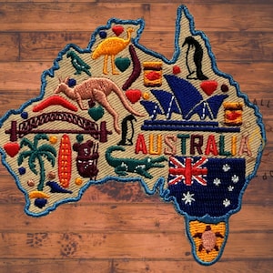 May include: Embroidered patch in the shape of Australia, featuring iconic symbols like a kangaroo, Sydney Opera House, and the Australian flag. The word "AUSTRALIA" is stitched across the centre. The patch is outlined in blue and set against a wood grain background.
