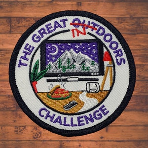 Puede incluir: Parche bordado con el texto "THE GREAT OUTDOORS IN CHALLENGE" en un diseño circular. El parche presenta una escena de montaña dentro de una pantalla de televisión, una lámpara roja y otros elementos temáticos al aire libre. El parche está sobre una superficie de madera.