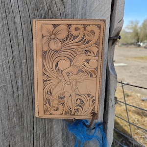 May include: A light brown leather wallet with a floral and cowboy design. The wallet features a detailed carving of a cowboy on a horse, surrounded by intricate floral patterns. The edges are stitched with black thread.