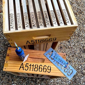 Puede incluir: Dos cajas de madera con el número A5118669 grabado en ellas. La caja superior tiene el número repetido en cada uno de los ocho lados. La caja inferior tiene el número grabado en la parte delantera y una plantilla de metal con el mismo número está encima de la caja.