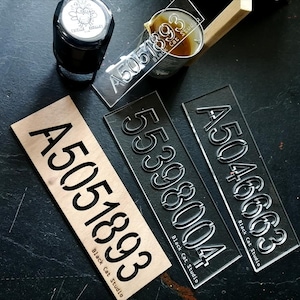 Op de afbeelding: Drie transparante acrylstencils met de nummers "A5051893", "A5046663" en "55398004" erop gedrukt. De stencils liggen op een donker oppervlak met een klein zwart inktkussen en een blikje zwarte verf.
