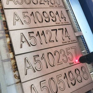 Op de afbeelding: Vijf houten plaquettes met lasergesneden nummers en de tekst "Black Cat Studio" op elke plaquette. De nummers op de plaquettes zijn A5062465, A5109914, A5112774, A5102357 en A5051988.