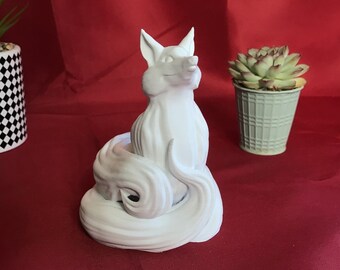 May include: A white fox figurine with a curled tail, sitting upright. The fox has pointed ears and a smiling expression. The figurine is on a red surface, with a checkered pot and a succulent plant in the background.