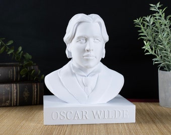 Może przedstawiać: Biała gipsowa popiersie Oscara Wilde'a na białej podstawie z napisem "Oscar Wilde" wygrawerowanym z przodu. Popiersie stoi na jasnobrązowej powierzchni tkanej.